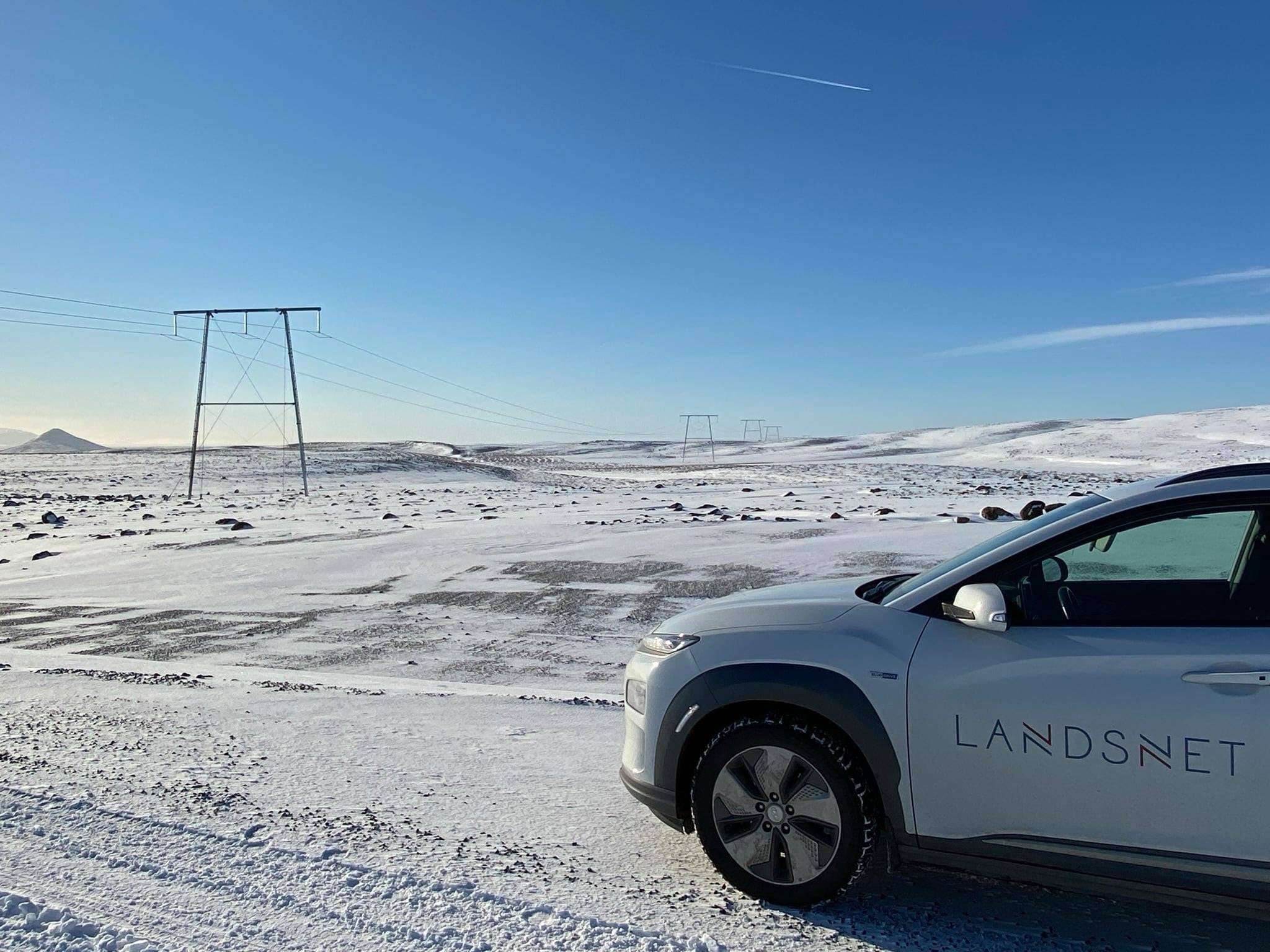 Rafmagnsbíll og mastur í snævi þökktu landslagi. Í loftinu má sjá rákir frá flugvél.